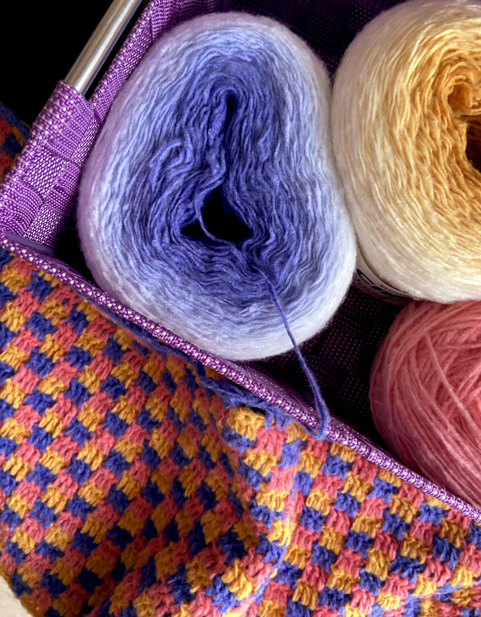 a checkerboard patterned crochet project lies next to a basket of working yarn